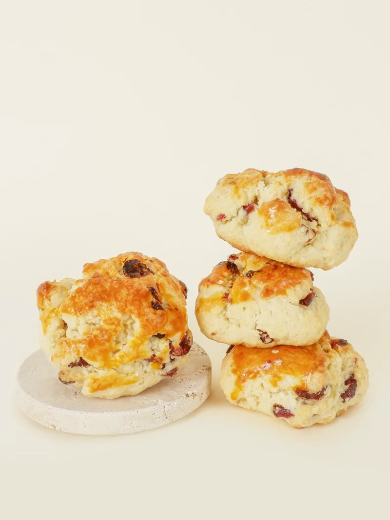 Four Cranberry scones on a cream backdrop, dried cranberries showing through the golden glaze — one on a stone coaster, three stacked.