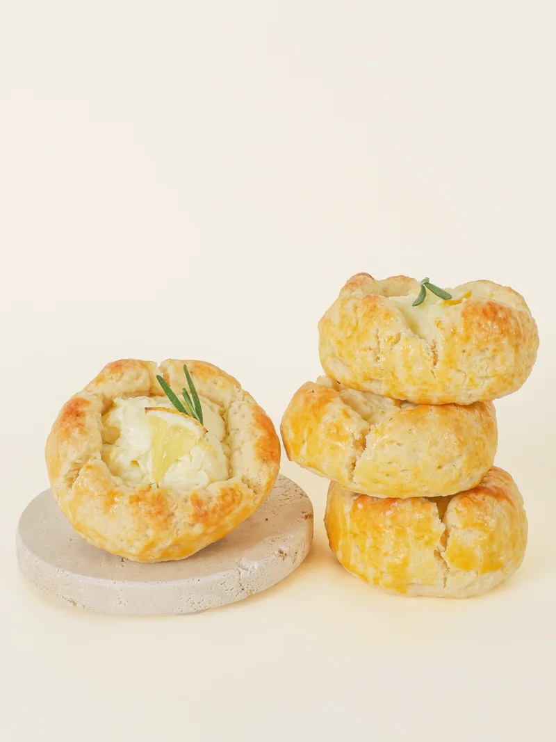 Four Lemon scones — one open on a stone coaster, its cream cheese centre topped with a slice of lemon and a sprig of rosemary, three stacked.