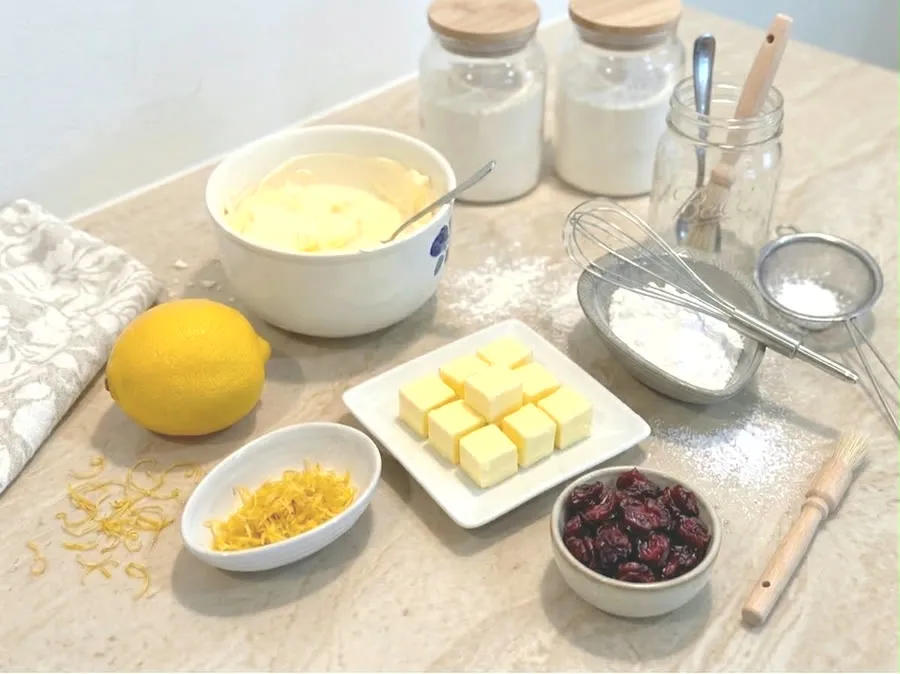 Ingredients set out separately on a marble counter — two jars of flour, cubes of butter on a plate, a bowl of cream cheese, dried cranberries, a lemon with its zest, and icing sugar beside a sieve and a whisk.